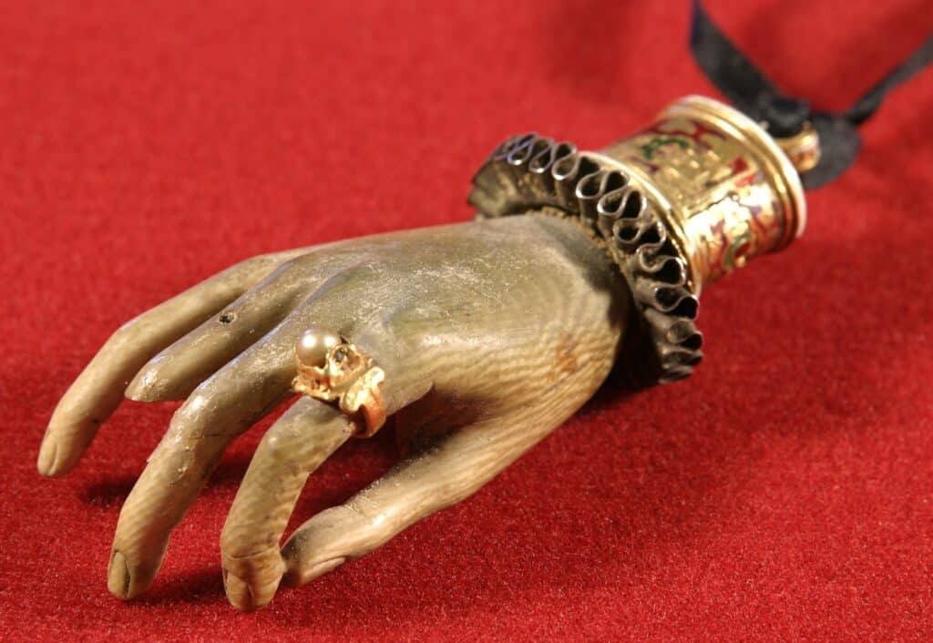 Exvot en forma de mà de plata daurada amb anell de perla, puny decorat amb esmalts i dentell metàl·lic, sobre fons de vellut vermell.