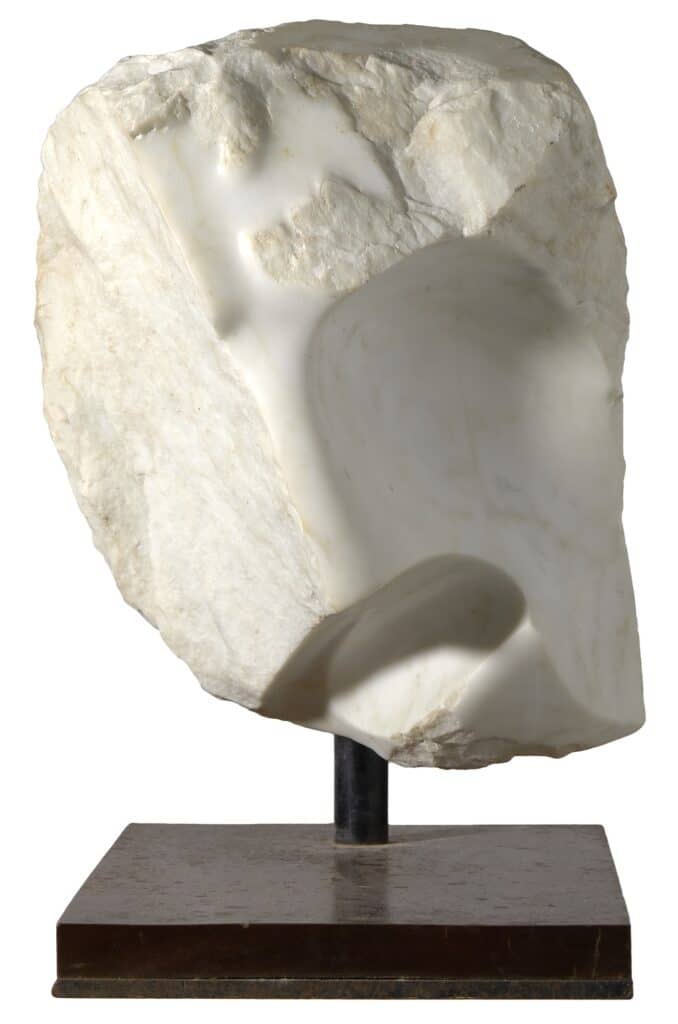 Escultura de marbre blanc parcialment desbastada amb un cap humà emergint de la pedra en brut, muntada sobre peanya de fusta.