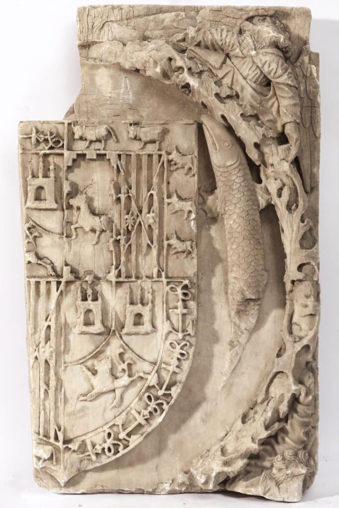 Fragment escultòric de pedra amb un escut heràldic decorat amb edificis, animals i barres, envoltat per un peix i ornamentació vegetal