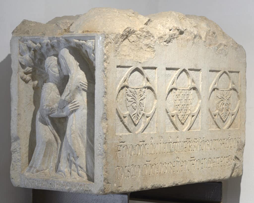 Fragment de pedra amb relleu gòtic: figures femenines besant-se a l'esquerra i tres escuts heràldics amb inscripció a la dreta