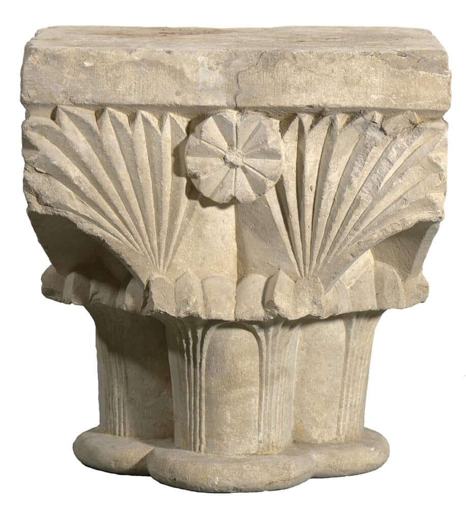 Capitell de pedra de forma cúbica amb decoració esculpida que combina palmetes i un motiu floral central. Les formes són simètriques i d’estil romànic, amb relleu marcat i línies netes. La pedra presenta desgast natural i to clar