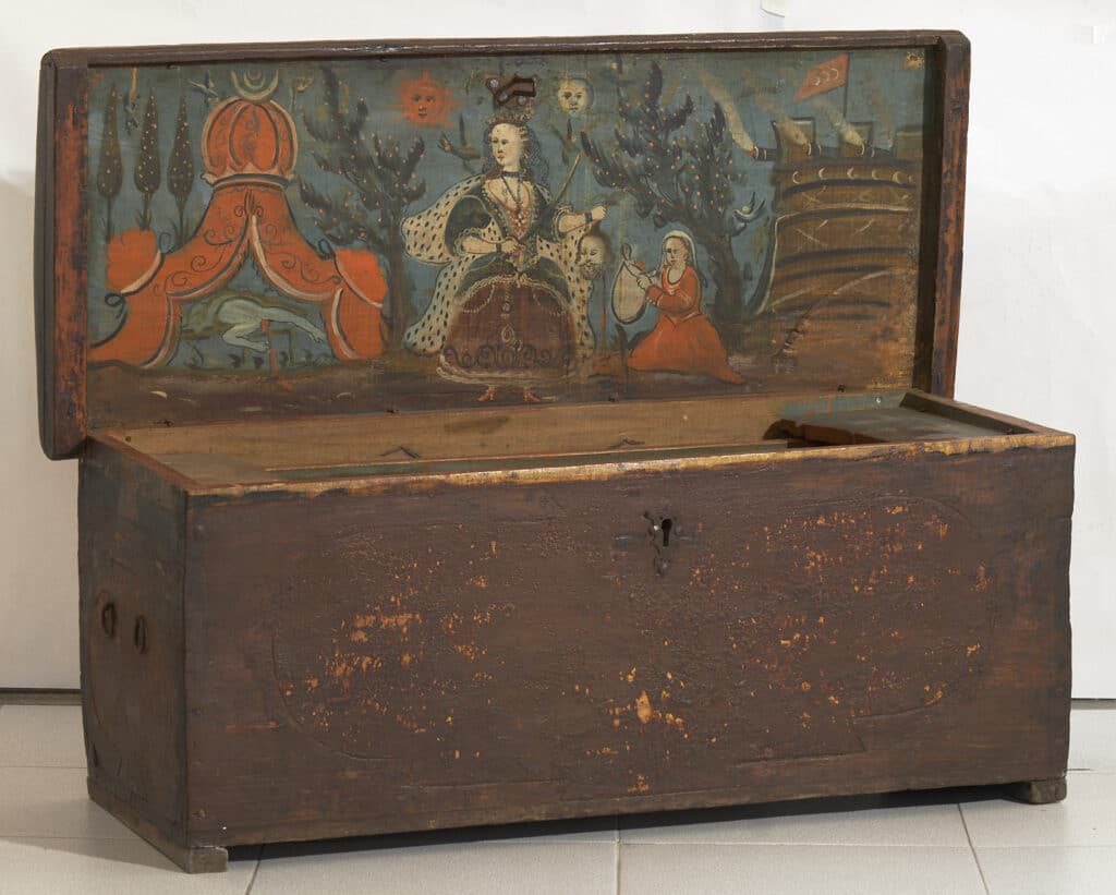 Arca de fusta antiga oberta amb interior pintat mostrant figura femenina amb vestit llarg entre elements decoratius sobre fons blau verdós.