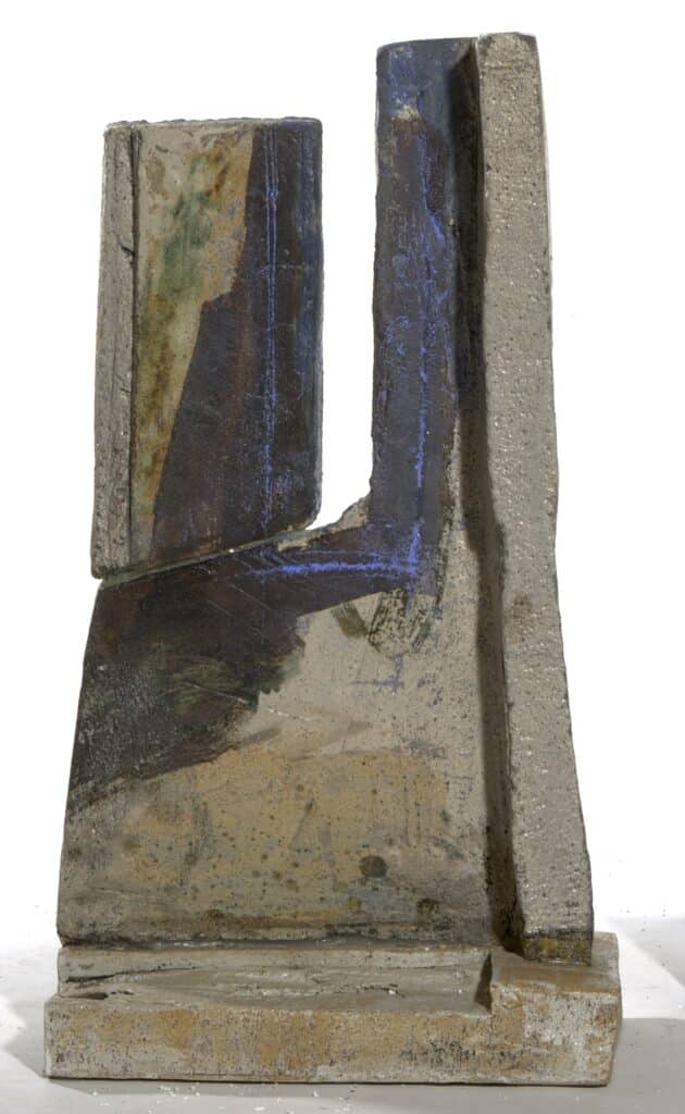 Escultura abstracta de formigó amb formes geomètriques verticals i traces de pintura blava, marró i verda sobre base quadrada