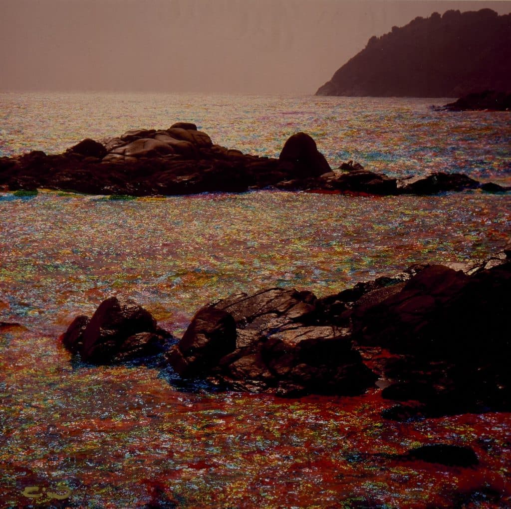 Aquesta imatge, titulada Palamòs de Tino Soriano, mostra roques fosques emergint del mar, amb l'aigua reflectint llums irisades en tons vermells, blaus, verds i grocs. Al fons, es veu un promontori difuminat en una atmosfera tranquil·la, amb una llum suau i càlida.