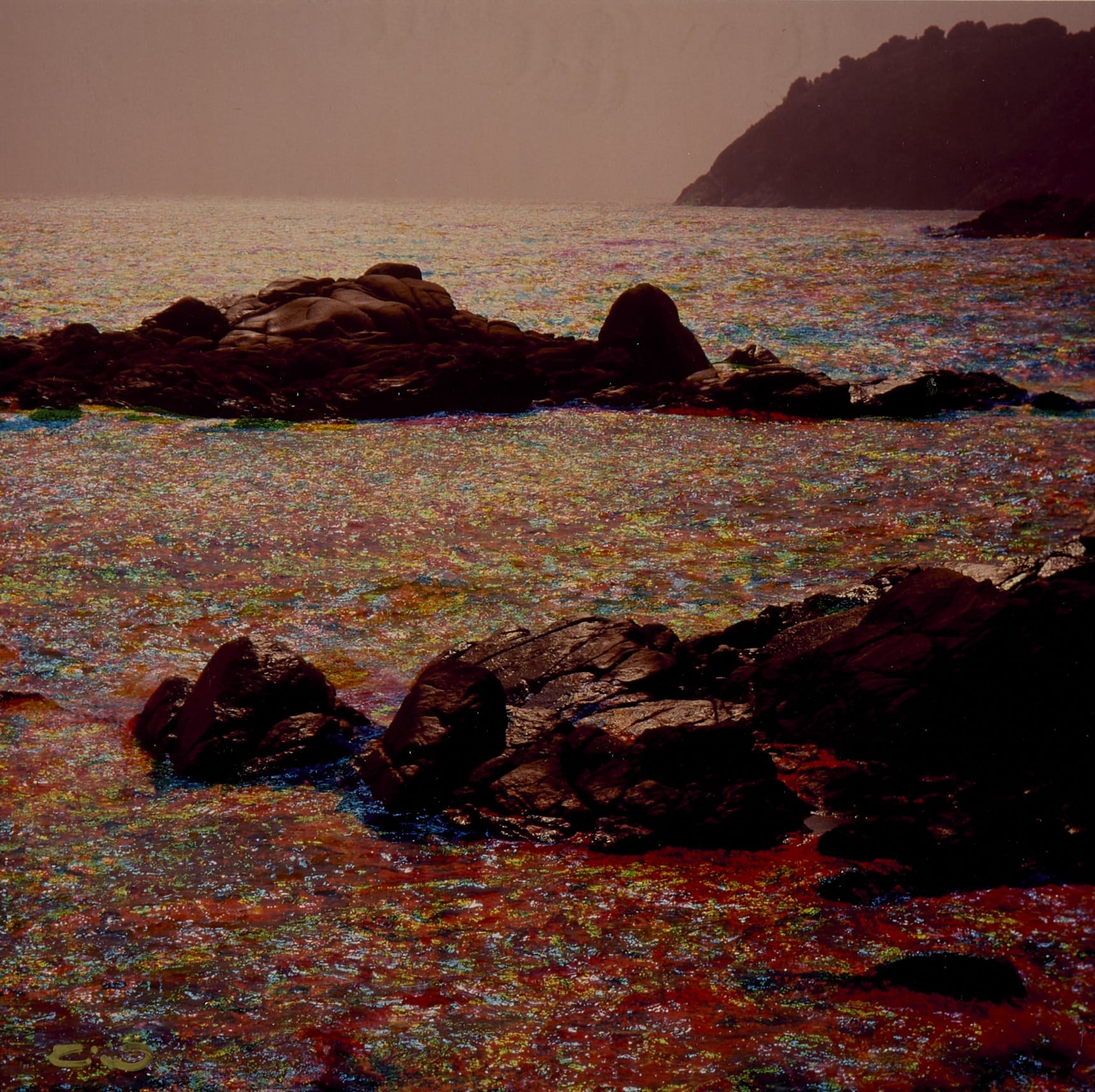 Aquesta imatge, titulada Palamòs de Tino Soriano, mostra roques fosques emergint del mar, amb l'aigua reflectint llums irisades en tons vermells, blaus, verds i grocs. Al fons, es veu un promontori difuminat en una atmosfera tranquil·la, amb una llum suau i càlida.