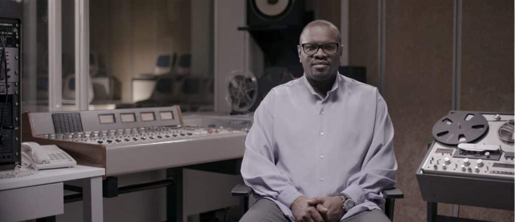 Home amb ulleres i camisa clara assegut en un estudi de gravació vintage amb taula de mescles analògica i magnetòfon de bobina oberta al fons